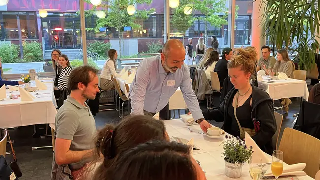 Murat Karakaya bringt einer Frau, die an einem Tisch sitzt, eine Speise. Die Atmosphäre ist gesellig, mit Gästen, die sich unterhalten. 
