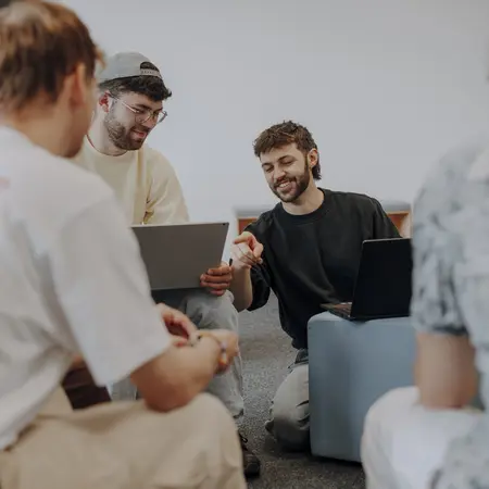 Man sieht fünf Studierende im Vordergrund, die gemeinsam an einem Projekt arbeiten und lächeln. Ein Studierender zeigt auf den Laptop des anderen und lacht dabei.