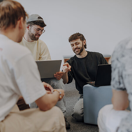 Man sieht fünf Studierende im Vordergrund, die gemeinsam an einem Projekt arbeiten und lächeln. Ein Studierender zeigt auf den Laptop des anderen und lacht dabei.