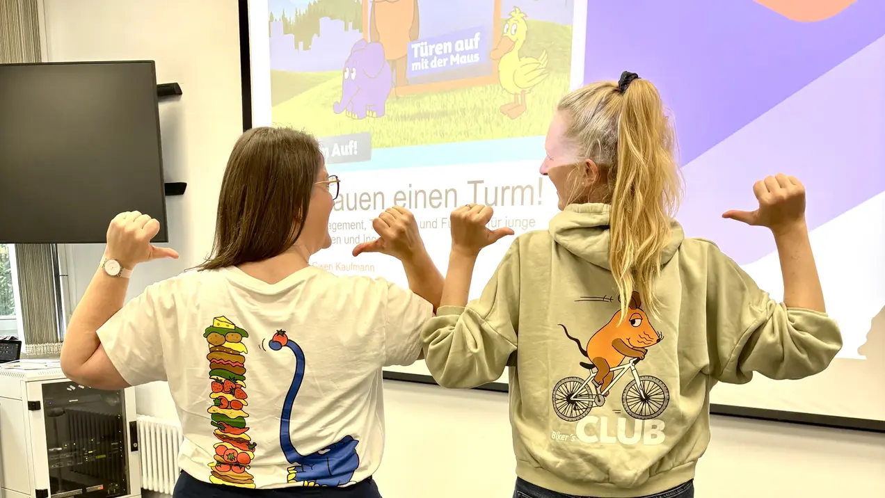 Zwei Personen stehen Rücken an Rücken in einem Raum. Die eine trägt ein T-Shirt mit bunten Mustern, die andere eine graue Jacke mit einem Tiermotiv. Im Hintergrund ist ein Bildschirm mit einer bunten Präsentation zu sehen.