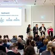 Eine Podiumsdiskussion zum Thema Mobilitätsmanagement in Stadt und Land. Fünf Personen stehen am Tisch, eine spricht ins Mikrofon. Hinter ihnen ist eine Präsentation mit dem Titel "Begrüßungstalk" und den Namen der Teilnehmenden. Im Vordergrund sind zahlreiche Zuhörer:innen zu sehen.