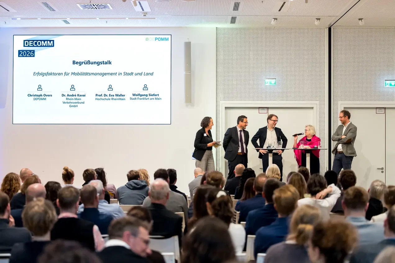 Eine Podiumsdiskussion zum Thema Mobilitätsmanagement in Stadt und Land. Fünf Personen stehen am Tisch, eine spricht ins Mikrofon. Hinter ihnen ist eine Präsentation mit dem Titel "Begrüßungstalk" und den Namen der Teilnehmenden. Im Vordergrund sind zahlreiche Zuhörer:innen zu sehen.
