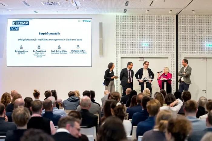 Eine Podiumsdiskussion zum Thema Mobilitätsmanagement in Stadt und Land. Fünf Personen stehen am Tisch, eine spricht ins Mikrofon. Hinter ihnen ist eine Präsentation mit dem Titel "Begrüßungstalk" und den Namen der Teilnehmenden. Im Vordergrund sind zahlreiche Zuhörer:innen zu sehen.