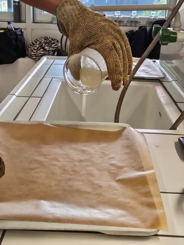 Eine Hand trägt einen hitzebeständigen Handschuh und gießt eine Flüssigkeit aus einem Glas in eine Form, die mit Backpapier ausgelegt ist. Im Hintergrund sind eine Spüle und verschiedene Utensilien zu sehen.
