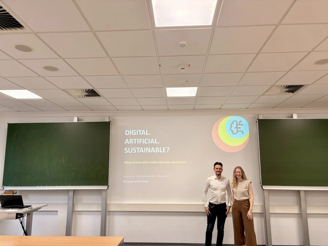 Ein Mann und eine Frau stehen in einem Seminarraum vor einer Präsentation mit dem Titel "DIGITAL. ARTIFICIAL. SUSTAINABLE?". Hintergrund: zwei grüne Tafeln und eine Tischplatte. Der Raum wirkt professionell und geeignet für eine akademische Veranstaltung.