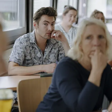 Man sieht Studierende, die gespannt nach vorne schauen und dem Kurs zuhören. Sie sitzen an Schreibtischen und ein Student insbesondere ist im Fokus.