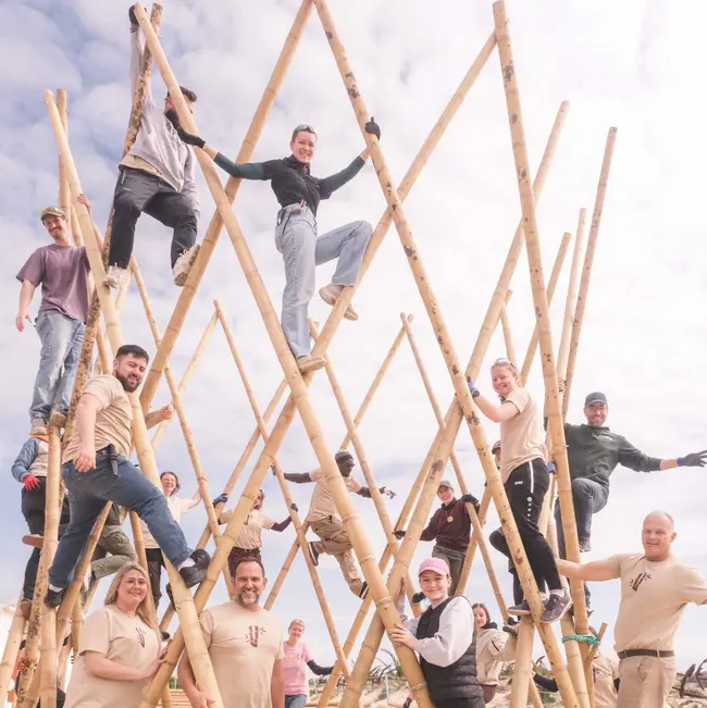Eine Gruppe von Menschen steht und sitzt auf einem großen, kunstvoll konstruierten Gerüst aus Bambusstangen. Die Teilnehmer lächeln und posieren, während sie verschiedene Posen einnehmen. Der Himmel ist bewölkt, und die Szene befindet sich im Freien.