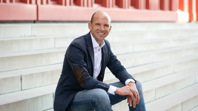 Ein lächelnder Mann sitzt auf einer Treppe. Er trägt ein Sakko und eine helle Hemd, und hat eine entspannte Körperhaltung. Der Hintergrund ist unscharf und zeigt architektonische Elemente.