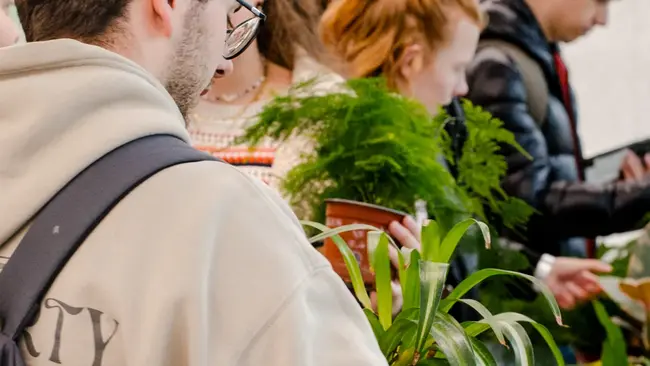 Eine Gruppe von Menschen steht an einem Tisch, der mit Pflanzen und Töpfen bedeckt ist. Ein junger Mann mit einer Brille hält eine Pflanze in der Hand. Andere Personen im Hintergrund schauen sich ebenfalls Pflanzen an.
