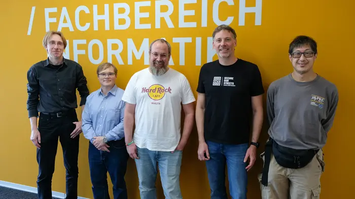 Fünf Personen stehen vor einer orangen Wand mit dem Schriftzug „Fachbereich Informatik“. Sie lächeln und posieren für das Foto. Die Männer tragen unterschiedliche Kleidungsstile, eine Person trägt ein T-Shirt mit dem Logo von Hard Rock.