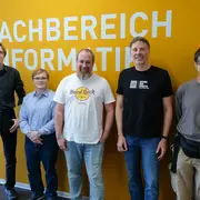 Fünf Personen stehen vor einer orangen Wand mit dem Schriftzug „Fachbereich Informatik“. Sie lächeln und posieren für das Foto. Die Männer tragen unterschiedliche Kleidungsstile, eine Person trägt ein T-Shirt mit dem Logo von Hard Rock.
