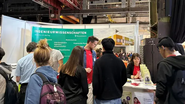 Eine Gruppe von Personen steht an einem Messestand, der Informationen über Ingenieurwissenschaften anbietet. Im Hintergrund sind Banner und ein Tisch mit Broschüren zu sehen. Ein Mitarbeiter spricht mit Besuchern, während eine weitere Person am Tisch sitzt und Informationen bereitstellt.