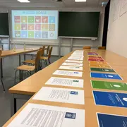 Auf einem Tisch liegen mehrere Informationsblätter zu den nachhaltigen Entwicklungszielen. Im Hintergrund ist eine Präsentation mit den Symbolen dieser Ziele sichtbar. Der Raum ist mit Stühlen und einer grünen Tafel ausgestattet.