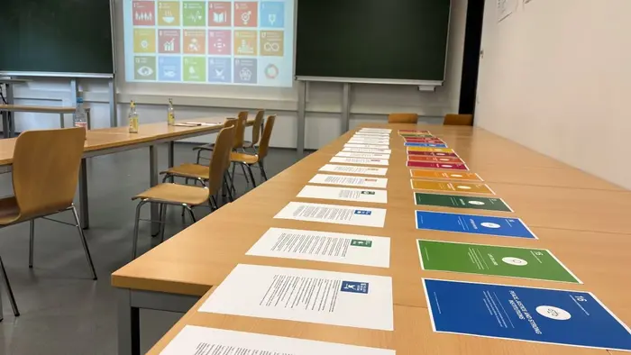 Auf einem Tisch liegen mehrere Informationsblätter zu den nachhaltigen Entwicklungszielen. Im Hintergrund ist eine Präsentation mit den Symbolen dieser Ziele sichtbar. Der Raum ist mit Stühlen und einer grünen Tafel ausgestattet.