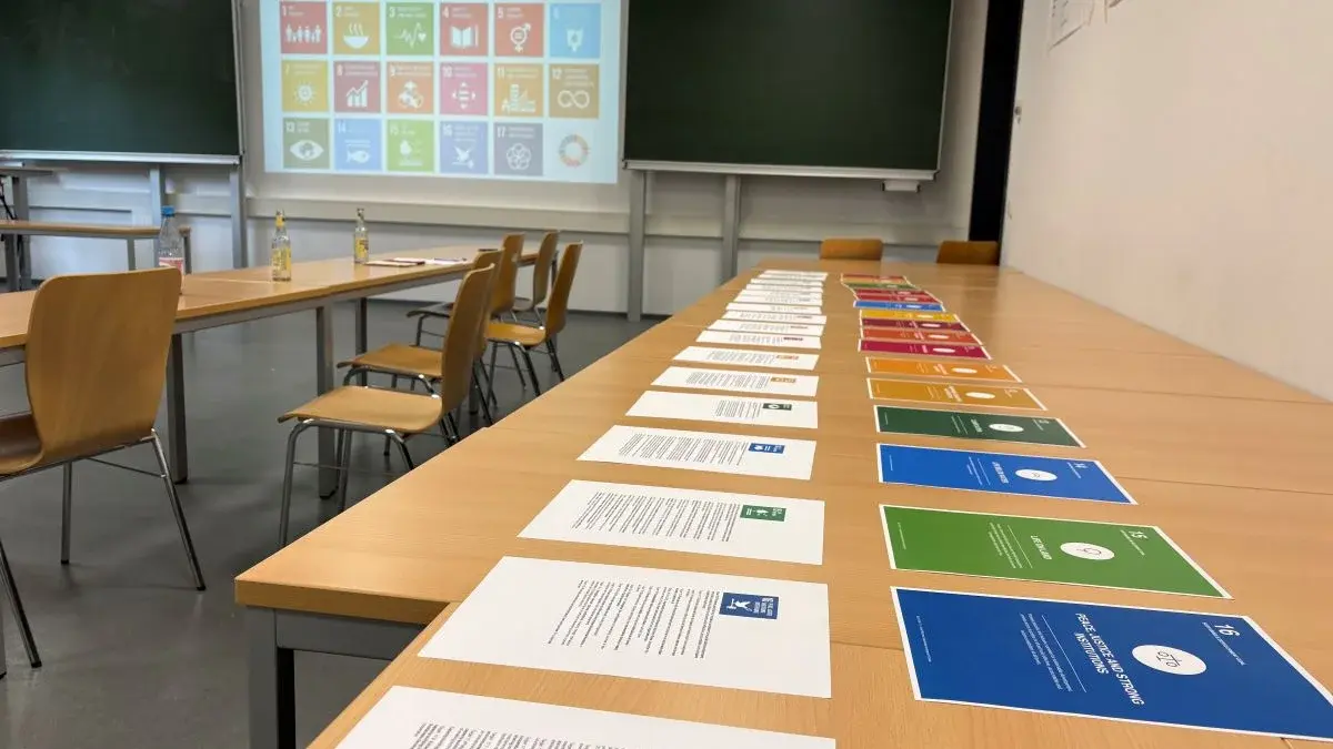 Auf einem Tisch liegen mehrere Informationsblätter zu den nachhaltigen Entwicklungszielen. Im Hintergrund ist eine Präsentation mit den Symbolen dieser Ziele sichtbar. Der Raum ist mit Stühlen und einer grünen Tafel ausgestattet.