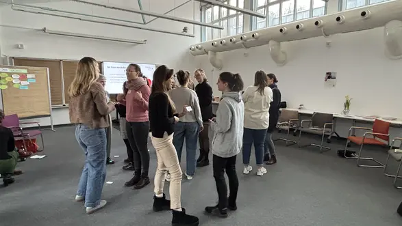 Eine Gruppe von Menschen steht in einem Raum und unterhält sich aktiv. Es gibt mehrere Teilnehmerinnen, die in informellen Kleidungen gekleidet sind. Im Hintergrund sind einige Stühle und eine Präsentationsleinwand zu sehen. Die Atmosphäre wirkt lebhaft und interaktiv.