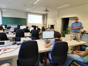 In einem Laborraum stehen mehrere Tische mit Computern und Messgeräten. Eine Gruppe von Studierenden arbeitet konzentriert daran, während ein Lehrer vorne steht und erklärt. Eine projizierte Präsentation ist an der Wand sichtbar.