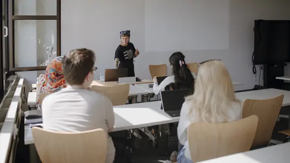 Man sieht eine Studierende, die einen Vortrag hält und einen Kurs, der ihr zuhört. Auf den Folien auf dem großen Bildschirm steht "Wie können wir es besser machen?"