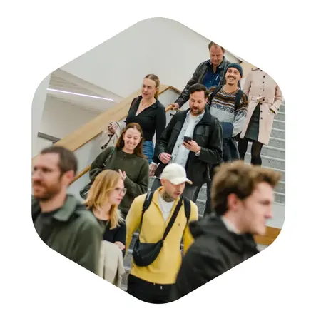 Eine Gruppe von Menschen geht eine Treppe hinunter. Die Personen tragen unterschiedliche Kleidung, einige schauen auf ihre Handys. Die Atmosphäre wirkt lebhaft und der Raum ist hell erleuchtet.