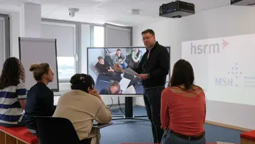 In einem modernen Seminarraum präsentiert ein Dozent vor einer Gruppe von fünf Personen. Auf dem großen Bildschirm sind zusätzliche Teilnehmer zu sehen, während die Zuschauer aufmerksam zuhören. Der Raum ist hell und für interaktive Schulungen eingerichtet.