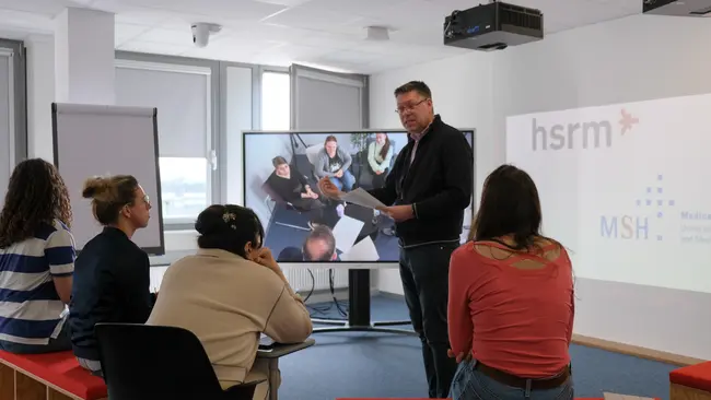 In einem modernen Seminarraum präsentiert ein Dozent vor einer Gruppe von fünf Personen. Auf dem großen Bildschirm sind zusätzliche Teilnehmer zu sehen, während die Zuschauer aufmerksam zuhören. Der Raum ist hell und für interaktive Schulungen eingerichtet.