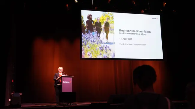Eine Frau steht am Rednerpult auf der Bühne und hält eine Präsentation. Im Hintergrund ist eine Folie mit der Aufschrift „Hochschule RheinMain – Ihre erste Anlaufstelle“ sowie das Datum und der Name des Vortragenden zu sehen. Der Raum ist beleuchtet, und das Publikum sitzt im Hintergrund.