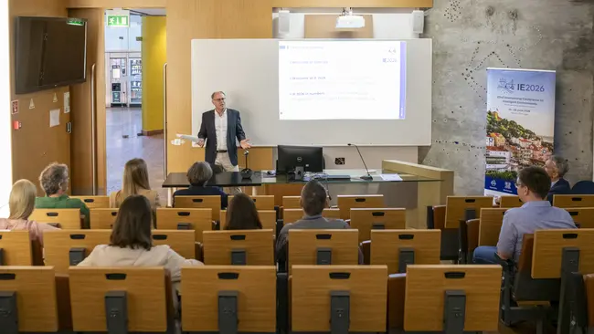 Ein Referent steht an einem Tisch und präsentiert vor einem Publikum in einem Seminarraum. Im Hintergrund ist eine Projektionsfläche mit Informationen zur Konferenz IE2026 zu sehen. Die Zuhörer sitzen in Reihen und hören aufmerksam zu.