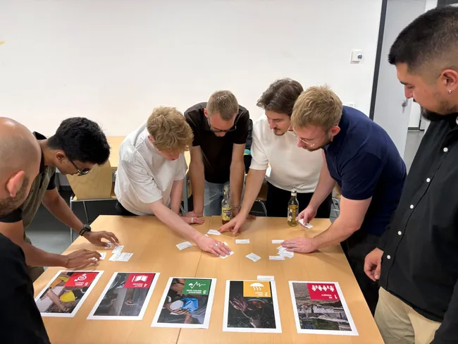 Eine Gruppe von sechs Personen steht um einen Tisch und diskutiert über verschiedene Karten oder Bilder. Auf dem Tisch sind mehrere Plakate mit unterschiedlichen Symbolen und Farben angeordnet. Die Teilnehmer scheinen aktiv an einer Aufgabenstellung zu arbeiten.