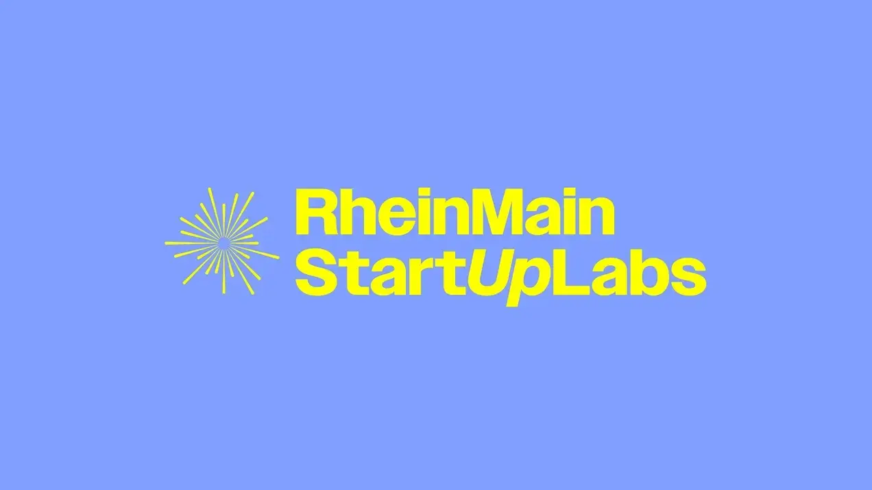 Das Bild zeigt den Schriftzug "RheinMain StartUpLabs" in gelber Schrift auf einem blauen Hintergrund. Neben dem Text befinden sich grafische Elemente, die an eine Sonne oder einen Stern erinnern.