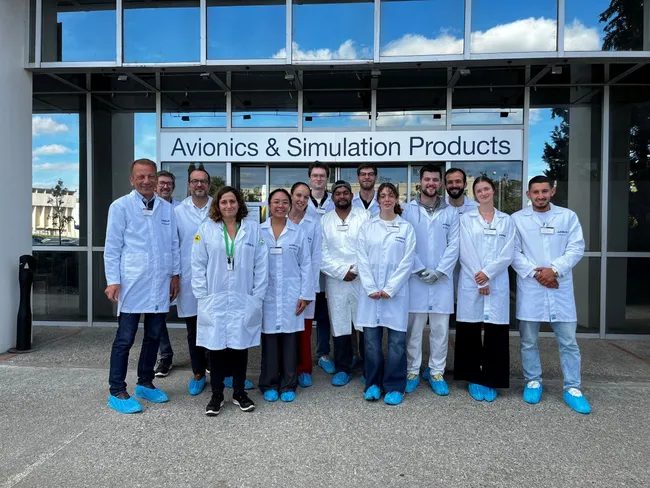 Eine Gruppe von zwölf Personen in weißen Laborkitteln steht vor einem Gebäude mit der Aufschrift "Avionics & Simulation Products". Einige tragen blaue Überziehschuhe. Die Personen lächeln und sehen freundlich aus.