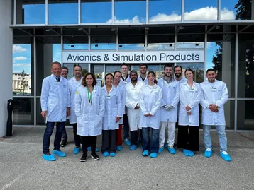 Eine Gruppe von zwölf Personen in weißen Laborkitteln steht vor einem Gebäude mit der Aufschrift "Avionics & Simulation Products". Einige tragen blaue Überziehschuhe. Die Personen lächeln und sehen freundlich aus.