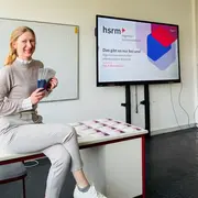 Eine Frau sitzt an einem Tisch und hält mehrere Prospekte in der Hand. Hinter ihr befindet sich ein Bildschirm mit der Aufschrift „hsrm Ingenieurwissenschaften“ und dem Slogan „Das gibt es nur bei uns!“. Die Atmosphäre wirkt freundlich und einladend.