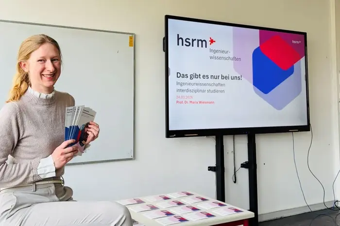 Eine Frau sitzt an einem Tisch und hält mehrere Prospekte in der Hand. Hinter ihr befindet sich ein Bildschirm mit der Aufschrift „hsrm Ingenieurwissenschaften“ und dem Slogan „Das gibt es nur bei uns!“. Die Atmosphäre wirkt freundlich und einladend.