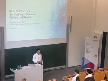Ein Referent steht an einem Rednerpult in einem Hörsaal und präsentiert über die Chancen, Risiken und Realität von KI im Studium und Berufsalltag. Im Hintergrund sind ein Bildschirm mit der Präsentation und eine Gruppe von Zuhörern zu sehen.