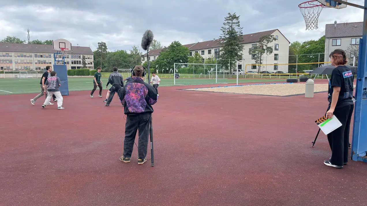 Eine Gruppe von Menschen spielt Basketball auf einem Sportplatz. Einige Personen sind mit Film- oder Aufnahmegeräten beschäftigt, während andere aktiv im Spiel sind. Im Hintergrund sind Volleyballnetze und Wohngebäude zu sehen. Der Himmel ist bewölkt.