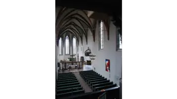 View of the interior of the church at Alsfeld Monastery with high vaulted ceilings and tall stained glass windows. In the foreground are rows of pews, with the altar area in the background. A modern work of art hangs on the wall. The atmosphere conveys a calm and sacred mood.
