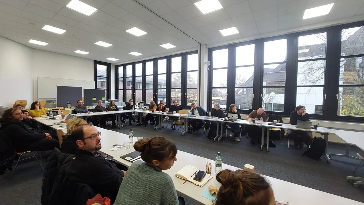 Menschen sitzen sich gegenüber in einer Art Kreis und lauschen einem Vortrag. 