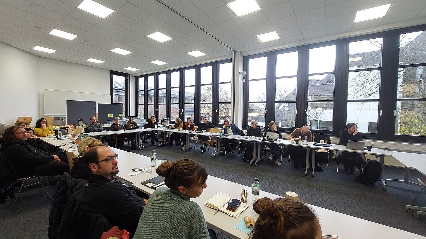 Menschen sitzen sich gegenüber in einer Art Kreis und lauschen einem Vortrag. 