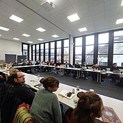 Menschen sitzen sich gegenüber in einer Art Kreis und lauschen einem Vortrag. 
