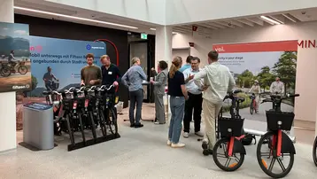 In einem modernen Raum sind mehrere Personen versammelt, die über Mobilität sprechen. Links stehen verschiedene Fahrräder, während rechts ein Informationsstand mit einem Banner zu sehen ist. Die Atmosphäre wirkt freundlich und informativ, geeignet für eine Veranstaltung zur nachhaltigen Fortbewegung.