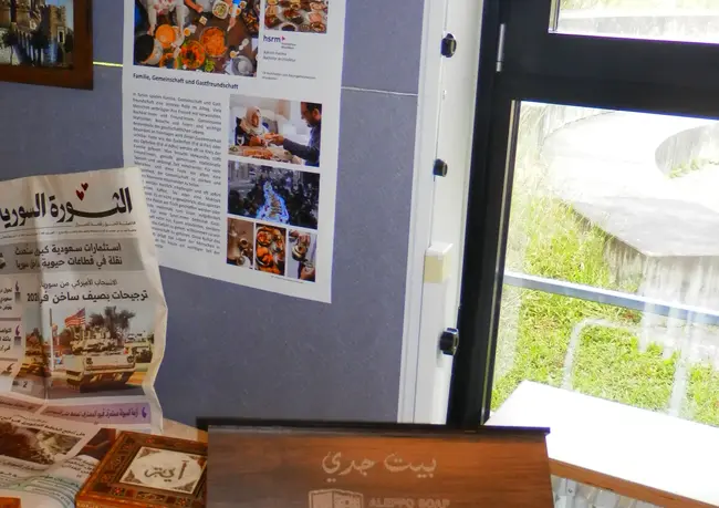 Ein Ausstellungsbereich zeigt verschiedene kunsthandwerkliche Gegenstände und Holzboxen auf einem Tisch. Im Hintergrund hängen informative Plakate über den arabischen Kulturstil. Auf einem Blatt Papier sind gedruckte Nachrichten sichtbar. Die Szenerie vermittelt ein Gefühl von kulturellem Austausch und traditionellem Handwerk.