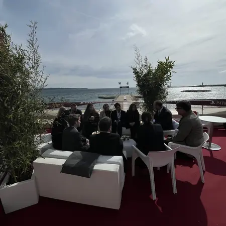 Eine Gruppe von Menschen sitzt auf weißen Sofas auf einer Terrasse mit Blick auf das Wasser. Die Umgebung ist mit Pflanzen dekoriert und die Stimmung wirkt entspannt. Im Hintergrund sind Boote und eine Küste sichtbar.