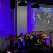 Eine Gruppe von Menschen, die in bunten Oberteilen zusammen singt, steht vor einem Klavier. Der Pianist sitzt mit dem Rücken zur Kamera. Im Hintergrund sind Lichter in lila und blauen Tönen zu sehen. Das Bild vermittelt eine fröhliche und musikalische Atmosphäre.