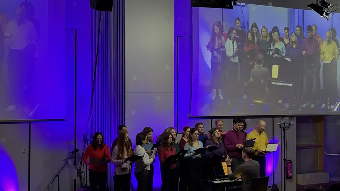 Eine Gruppe von Menschen, die in bunten Oberteilen zusammen singt, steht vor einem Klavier. Der Pianist sitzt mit dem Rücken zur Kamera. Im Hintergrund sind Lichter in lila und blauen Tönen zu sehen. Das Bild vermittelt eine fröhliche und musikalische Atmosphäre.