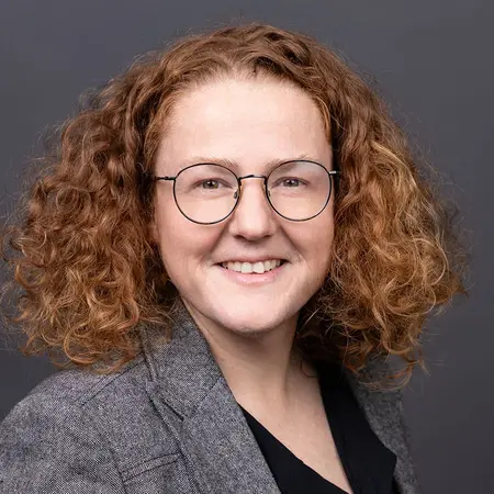 Ein lächelnde Frau mit lockigem, kupferfarbenem Haar trägt eine Brille und einen grauen Blazer. Sie steht vor einem neutralen, dunklen Hintergrund und vermittelt einen freundlichen und professionellen Eindruck.