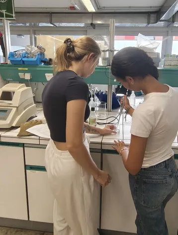 Zwei Frauen stehen in einem Labor und arbeiten an einer wissenschaftlichen Untersuchung. Die eine hält eine Pipette, während die andere Notizen macht. Es sind verschiedene Laborgeräte und Materialien im Hintergrund zu sehen.