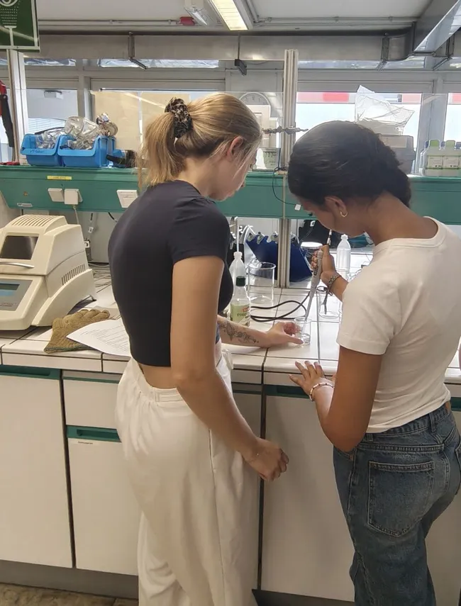 Zwei Frauen stehen in einem Labor und arbeiten an einer wissenschaftlichen Untersuchung. Die eine hält eine Pipette, während die andere Notizen macht. Es sind verschiedene Laborgeräte und Materialien im Hintergrund zu sehen.