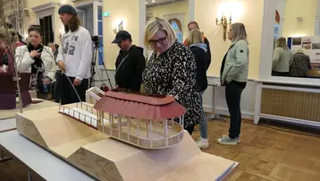 Eine Gruppe von Menschen steht in einem Ausstellungsraum. Im Vordergrund betrachtet eine Frau ein Modell eines Gebäudes mit einem roten Dach. Weitere Besucher sind im Hintergrund zu sehen, die ebenfalls mit dem Modell interagieren.