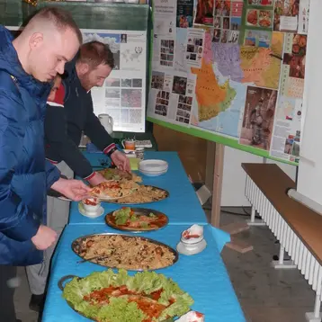 Zwei Personen stehen an einem Tisch und servieren sich Essen von mehreren großen Platten mit verschiedenen Gerichten, darunter Pizza und Salate. Im Hintergrund ist eine Wandtafel mit Landkarten und Informationen sichtbar. Die Umgebung wirkt festlich.