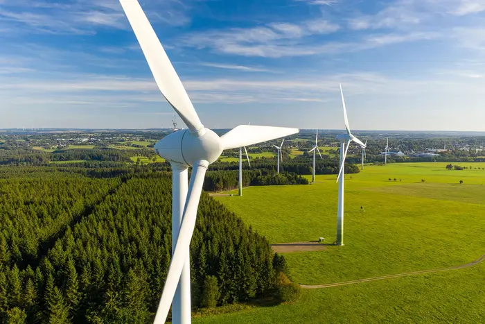 Luftaufnahme eines Windparks mit mehreren Windturbinen inmitten von grünen Wiesen und Wäldern. Der Himmel ist blau mit einigen Wolken, und die Landschaft erstreckt sich weitläufig in den Hintergrund.